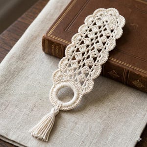 May include: Cream-colored, crocheted bookmark with a scalloped edge and a tassel. The bookmark features an openwork design and a circular loop at the bottom. It is resting on a linen surface next to a closed, brown book.