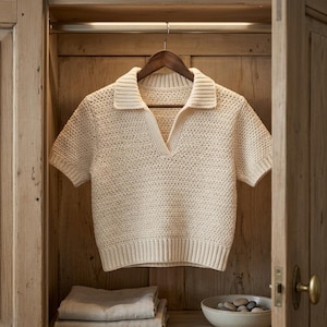 May include: Cream-colored crochet polo shirt with short sleeves, hanging on a wooden hanger inside a wooden wardrobe. The shirt features a collared neckline and a V-neck opening. The texture of the crochet is visible, and the shirt appears to be handmade.