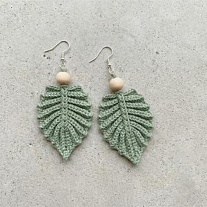 May include: A pair of handmade, sage green crochet leaf earrings. Each earring features a leaf shape with detailed stitching, a wooden bead, and a silver-colored hook. The earrings are displayed on a light gray surface.