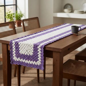 May include: A handmade crochet table runner in white and purple stripes. The runner is placed on a wooden table, with a cup and spoon on top. The runner's design features a granny square pattern at the ends.