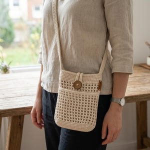 May include: A cream-coloured crochet crossbody bag with a long strap. The bag features a wooden button closure and a small pocket on the front. The bag is made of a woven, open-weave design.