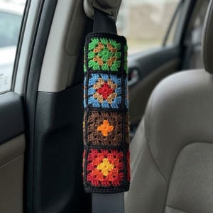 May include: A rectangular seat belt cover featuring a series of four stacked crochet granny squares. The squares are crafted in vibrant colors including green, blue, brown, orange, yellow, and red, all bordered by black yarn stitching.