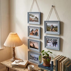 Può includere: Due set di cornici per foto sospese con bordi grigi e corda naturale. Ogni set contiene tre foto. Le cornici sono montate su una parete di colore chiaro. Una panca di legno con una lampada, libri e una pianta è in primo piano.