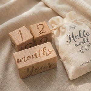 May include: A set of wooden baby milestone blocks with engraved numbers and text. The blocks show the numbers 1 and 2, plus the words "months" and "Year". A small drawstring bag with the words "Hello world" is also included.