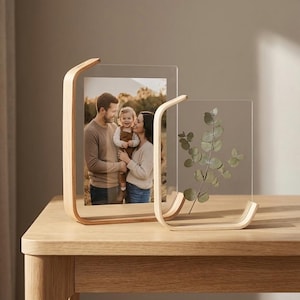 Peut inclure: Deux cadres photo en acrylique transparent avec des supports en bois incurvés. L'un contient une photo de famille, l'autre une branche d'eucalyptus. Les cadres sont posés sur une table en bois clair.