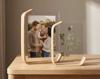 Könnte beinhalten: Zwei transparente Acryl-Fotorahmen mit gebogenen Holzständern. Ein Rahmen enthält ein Familienfoto, der andere einen Eukalyptuszweig. Die Rahmen stehen auf einem hellen Holztisch.