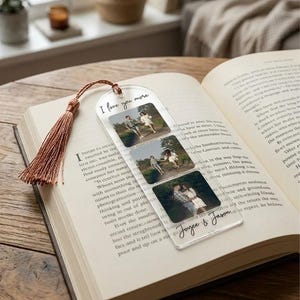 Peut inclure: Marque-page en acrylique transparent avec trois encarts photo et un pompon marron. Le marque-page indique "I love you more" et "Joyce & Jason". Le marque-page est placé dans un livre ouvert.