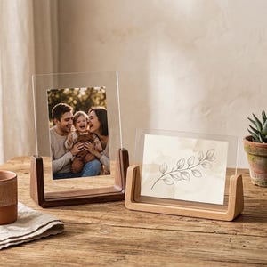 Könnte beinhalten: Zwei transparente Acryl-Fotorahmen mit Holzsockeln. Ein Rahmen enthält ein Familienfoto, der andere eine botanische Illustration. Die Rahmen stehen auf einer Holzoberfläche, mit einer kleinen Topfpflanze im Hintergrund.
