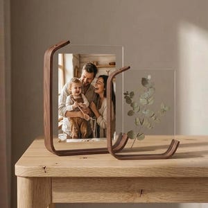 Op de afbeelding: Twee heldere acryl fotolijsten met gebogen walnoothouten steunen. Eén lijst bevat een familiefoto, de andere een takje eucalyptus. De lijsten staan op een lichte houten tafel, wat een moderne, minimalistische esthetiek creëert.