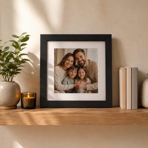 Peut inclure: Un cadre photo carré noir contenant une photographie couleur d'une famille de quatre personnes, deux adultes et deux enfants, portant tous des hauts à manches longues de tons neutres. Le cadre présente une large bordure blanche autour de l'image.