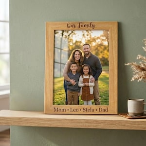 Peut inclure: Un cadre photo en bois clair avec une photo de famille. Le cadre porte l'inscription "Our Family" en haut et les noms "Mom • Leo • Stela • Dad" en bas. Le cadre est posé sur une étagère en bois.
