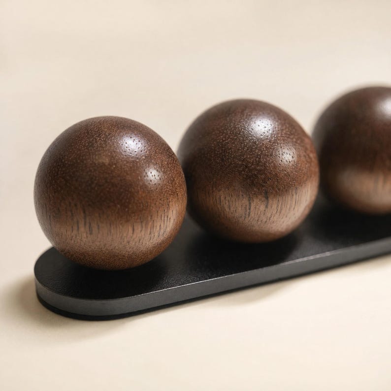 May include: Three smooth, spherical dark brown wooden balls are mounted in a linear arrangement on a flat, elongated black base. The wood grain is visible on the surface of each sphere, which reflects light, creating a highlight on the top.