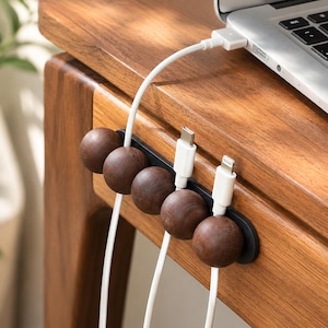 May include: A black cable organizer strip mounted on a wooden surface, featuring five dark brown spherical wooden magnetic clips that hold white charging cables in place, with one cable connected to a silver laptop on the desk.