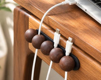May include: A black cable organizer strip mounted on a wooden surface, featuring five dark brown spherical wooden magnetic clips that hold white charging cables in place, with one cable connected to a silver laptop on the desk.