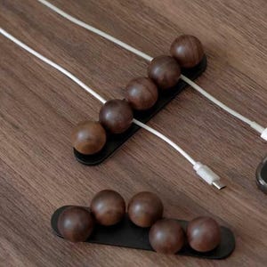 May include: Two black rectangular cable organizers featuring dark brown wooden spheres arranged in a row. The top organizer holds white charging cables between the spheres, while the bottom organizer is empty, showcasing the smooth wooden ball design.