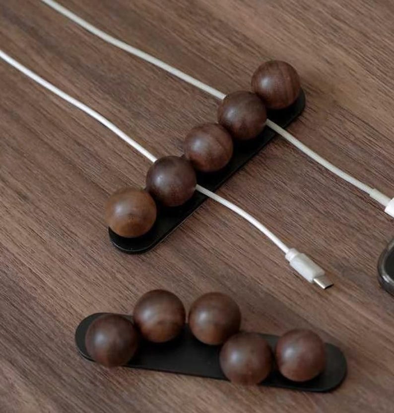 May include: Two black rectangular cable organizers featuring dark brown wooden spheres arranged in a row. The top organizer holds white charging cables between the spheres, while the bottom organizer is empty, showcasing the smooth wooden ball design.