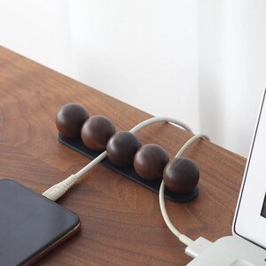May include: A cable organizer featuring five dark brown wooden spheres mounted on a black rectangular base. The device holds two white charging cables in place, with one cable connected to a smartphone and the other to a laptop computer.