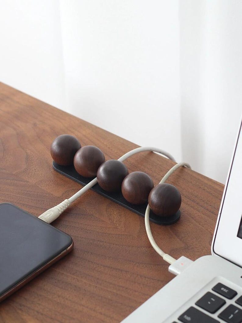 May include: A cable organizer featuring five dark brown wooden spheres mounted on a black rectangular base. The device holds two white charging cables in place, with one cable connected to a smartphone and the other to a laptop computer.