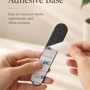 May include: A narrow, elongated strip with a dark gray felt-like top surface and a white adhesive backing labeled with 3M 9448A Double Coated Tissue Tape branding. The white protective film is partially peeled back to reveal the sticky adhesive layer.