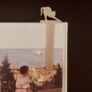 May include: A gold-colored metal bookmark in the shape of a cat, arched and stretching. The bookmark is inserted into a book with a painting of a woman overlooking a coastal town. The bookmark is approximately 6 inches long.