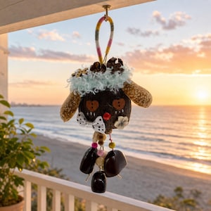 May include: A hanging ornament crafted from a dark brown coconut shell featuring two brown button eyes, a small red nose, and a white polka-dot bow tie. It is adorned with light blue fuzzy trim, wooden accents, and dangling dark brown seed pods.