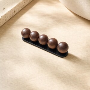 May include: A wall-mounted hook rack featuring five dark brown wooden spheres attached to a slim, horizontal black metal base. The spheres are arranged in a straight line, providing a functional design for hanging items on a flat surface.