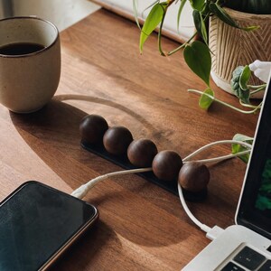 May include: A black cable organizer featuring five dark brown wooden spheres arranged in a row on a rectangular base. The device is positioned on a wooden surface, with white charging cables threaded between the spheres to connect a smartphone and a laptop.
