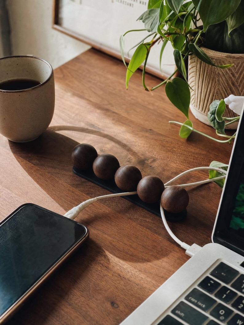 May include: A black cable organizer featuring five dark brown wooden spheres arranged in a row on a rectangular base. The device is positioned on a wooden surface, with white charging cables threaded between the spheres to connect a smartphone and a laptop.