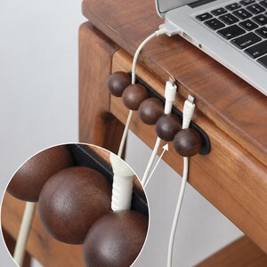 May include: A dark brown wooden cable organizer featuring five spherical beads mounted on a black base. The organizer is attached to the edge of a wooden desk, holding two white charging cables in place while a laptop sits on the desk surface above.