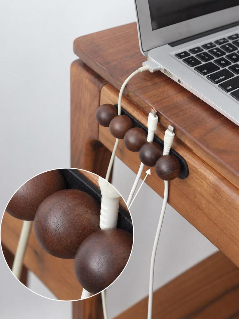 May include: A dark brown wooden cable organizer featuring five spherical beads mounted on a black base. The organizer is attached to the edge of a wooden desk, holding two white charging cables in place while a laptop sits on the desk surface above.
