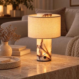 May include: A table lamp featuring a cylindrical base made of white stone with dark brown veining. The lamp has a circular, light-colored fabric shade and a small circular power button on the front of the base, along with a USB-C charging port.