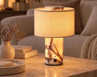 Può includere: Una lampada da tavolo con base cilindrica in pietra bianca con venature marrone scuro. La lampada presenta un paralume circolare in tessuto di colore chiaro, un piccolo pulsante di accensione circolare sulla base e una porta di ricarica USB-C.