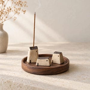 May include: A circular dark brown wooden tray holding three small, textured, beige ceramic house-shaped incense holders with dark brown roofs. One holder contains a single lit incense stick with a thin trail of smoke rising upward.