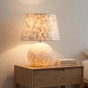 May include: A table lamp featuring a spherical base covered in small, iridescent square tiles and a tapered lampshade adorned with overlapping, translucent circular shell-like pieces that emit a warm, soft glow when illuminated.