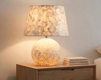 Può includere: Una lampada da tavolo con base sferica rivestita di piccole tessere quadrate iridescenti e un paralume conico decorato con elementi circolari traslucidi sovrapposti, simili a conchiglie, che emettono una luce calda e soffusa.