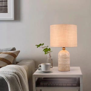 Pu&ograve; includere: Una lampada da tavolo con base cilindrica in pietra beige testurizzata e paralume a tamburo in tessuto color marrone chiaro. La lampada emette una luce calda e poggia su un tavolino bianco accanto a una tazza in ceramica e un vaso in vetro.
