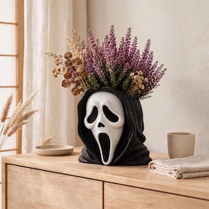 May include: A ceramic vase shaped like a white ghost mask with a black hooded shroud, filled with a bouquet of purple heather, dried brown eucalyptus leaves, and small beige flowers, creating a decorative container for floral arrangements.