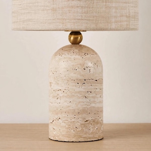 Pu&ograve; includere: Una lampada da tavolo con una base cilindrica in pietra travertino beige porosa, caratterizzata da una texture naturale irregolare. La lampada presenta un piccolo elemento di giunzione sferico liscio color ottone e un ampio paralume cilindrico in tessuto color sabbia.