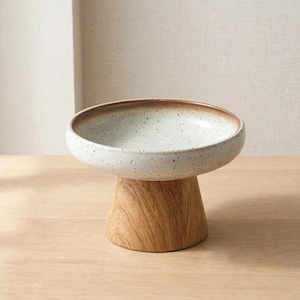 以下が含まれることがあります： 茶色の縁取りが施された、斑点のあるオフホワイトの陶器製ボウル。広がりのある明るい木製の台座に支えられています。ボウルは幅広で浅い形状をしており、滑らかで丸みを帯びた縁と天然木の木目が特徴的です。