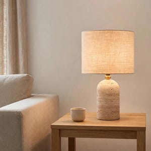 May include: A table lamp with a cylindrical, porous beige stone base and a gold-toned metal neck. The lamp features a drum-shaped, light-colored fabric shade that emits a warm glow. A small, matching beige stone cup sits beside the lamp base.