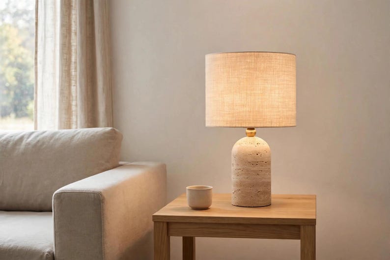 Pu&ograve; includere: Una lampada da tavolo con base cilindrica in pietra beige porosa e collo in metallo dorato. La lampada presenta un paralume in tessuto chiaro a forma di tamburo che emette una luce calda. Una piccola tazza in pietra beige abbinata &egrave; accanto.