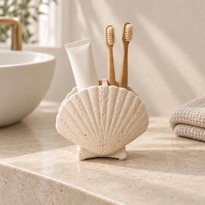 May include: A beige, textured seashell-shaped holder designed for bathroom storage. The holder contains two wooden-handled toothbrushes with light-colored bristles and one plain white, unmarked tube of toothpaste standing upright inside.