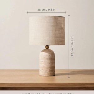 Pu&ograve; includere: Una lampada da tavolo in travertino con base cilindrica in pietra di travertino naturale, un dettaglio in metallo color ottone e un paralume a tamburo in lino naturale. La lampada misura 42 cm di altezza e 25 cm di diametro, con attacco E27.