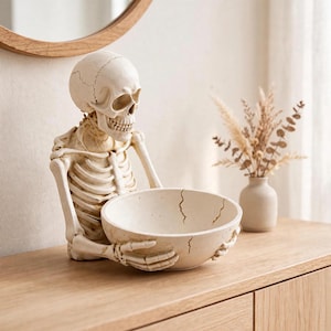 May include: A cream-coloured skeleton figurine holding a matching round bowl with a cracked design. The skeleton features a detailed skull, rib cage, and arms, while the bowl is positioned in its hands, creating a functional decorative piece.
