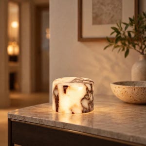 May include: A cylindrical table lamp crafted from polished stone with a translucent quality, featuring prominent dark brown and black veining patterns against a creamy white base, emitting a warm, soft glow from within its solid structure.