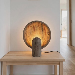 Pode incluir: Um candeeiro de mesa circular com uma base de pedra cinzenta texturizada e uma moldura circular a condizer. O interior da moldura é iluminado por uma luz quente e dourada, realçando os padrões naturais salpicados de castanho e bege.