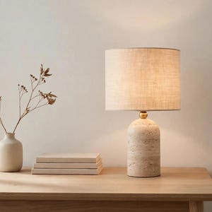 Pu&ograve; includere: Una lampada da tavolo con base cilindrica in pietra travertino, un piccolo connettore sferico dorato e un paralume a tamburo in tessuto beige. La lampada emette una luce soffusa ed &egrave; posizionata accanto a tre libri e un piccolo vaso in ceramica.