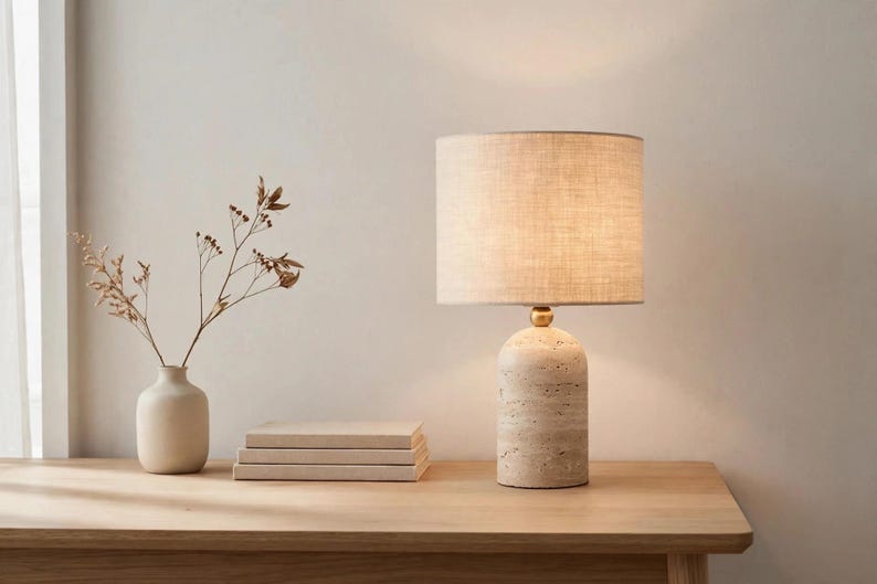 Pu&ograve; includere: Una lampada da tavolo con base cilindrica in pietra travertino, un piccolo connettore sferico dorato e un paralume a tamburo in tessuto beige. La lampada emette una luce soffusa ed &egrave; posizionata accanto a tre libri e un piccolo vaso in ceramica.