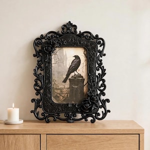 May include: A rectangular black picture frame with ornate scrollwork and sculpted rose accents. The frame contains an illustration of a black crow perched on a stone pillar against a backdrop of bare trees and a pale sky, with a white candle nearby.