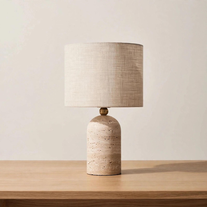 Pu&ograve; includere: Una lampada da tavolo con base cilindrica in pietra travertino beige porosa con parte superiore liscia e arrotondata. Una piccola sfera color ottone collega la base a un paralume cilindrico rivestito in tessuto beige chiaro testurizzato.